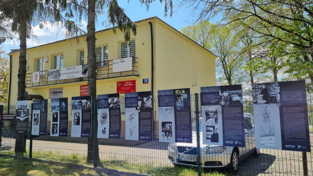 Widok na wystawę "Lem nie z tej ziemi" zamontowanej na ogrodzeniu wokół budynku Ochotniczej Straży Pożarnej w Puławach przy ul. Włodowickiej 36. W tle budynek Straży.