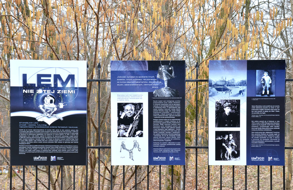 3 plansze wystawy "Lem nie z tej ziemi" zamontowane na ogrodzeniu. Na planszach fotografie przedstawiające Stanisława Lema, grafiki i tekst. W tle pożółkłe liście na ziemi i kwitnąca leszczyna.