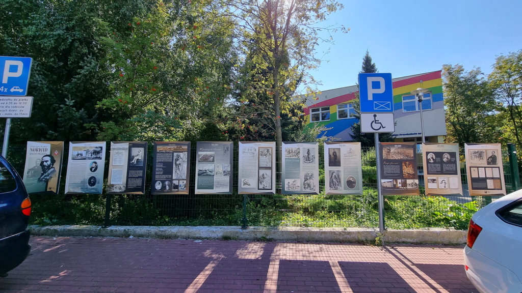 Widok na wystawę "Norwid wieczny pielgrzym" zamontowaną na ogrodzeniu Miejskiego Przedszkola nr 3 w Puławach przy ul. Legionu Puławskiego 8. W tle drzewa i budynek przedszkola.