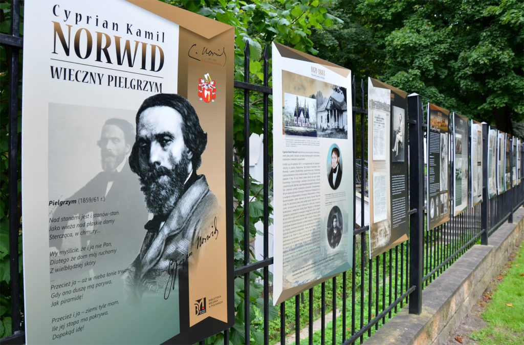 Widok na wystawę "Norwid wieczny pielgrzym" zamontowaną na ogrodzeniu wokół Biblioteki Miejskiej w Puławach przy ul. Głębokiej 7A. 