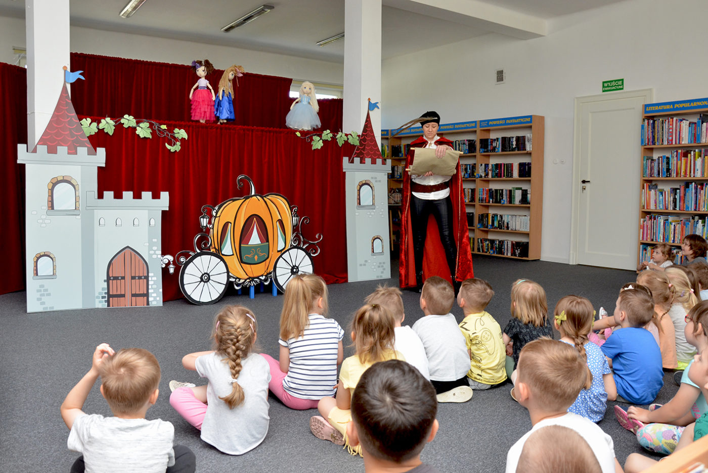 Zdjęcie z przedstawienia "Kopciuszek" zorganizowanego z okazji Dnia Postaci z bajek. Na zdjęcie pomieszczenie biblioteczne. na podłodze siedzą dzieci przodem do scenografii. scenografia: Między kolumnami rozpostarta bordowa kurtyna, przed nia mury zamku i kareta z dyni, powyżej 3 lalki. Przed dziećmi stoi osoba przebrana na herolda.