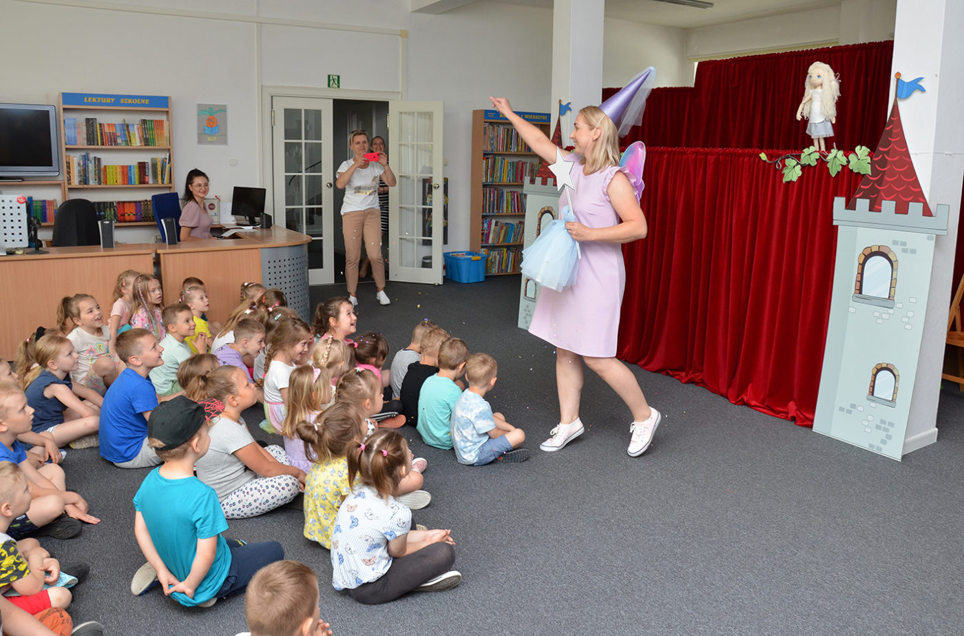 Na zdjęciu: pomieszczenie biblioteczne. Na podłodze siedzą dzieci przodem do sceny. Przed sceną osoba przebrana za wróżkę posypuje dzieci konfetti.