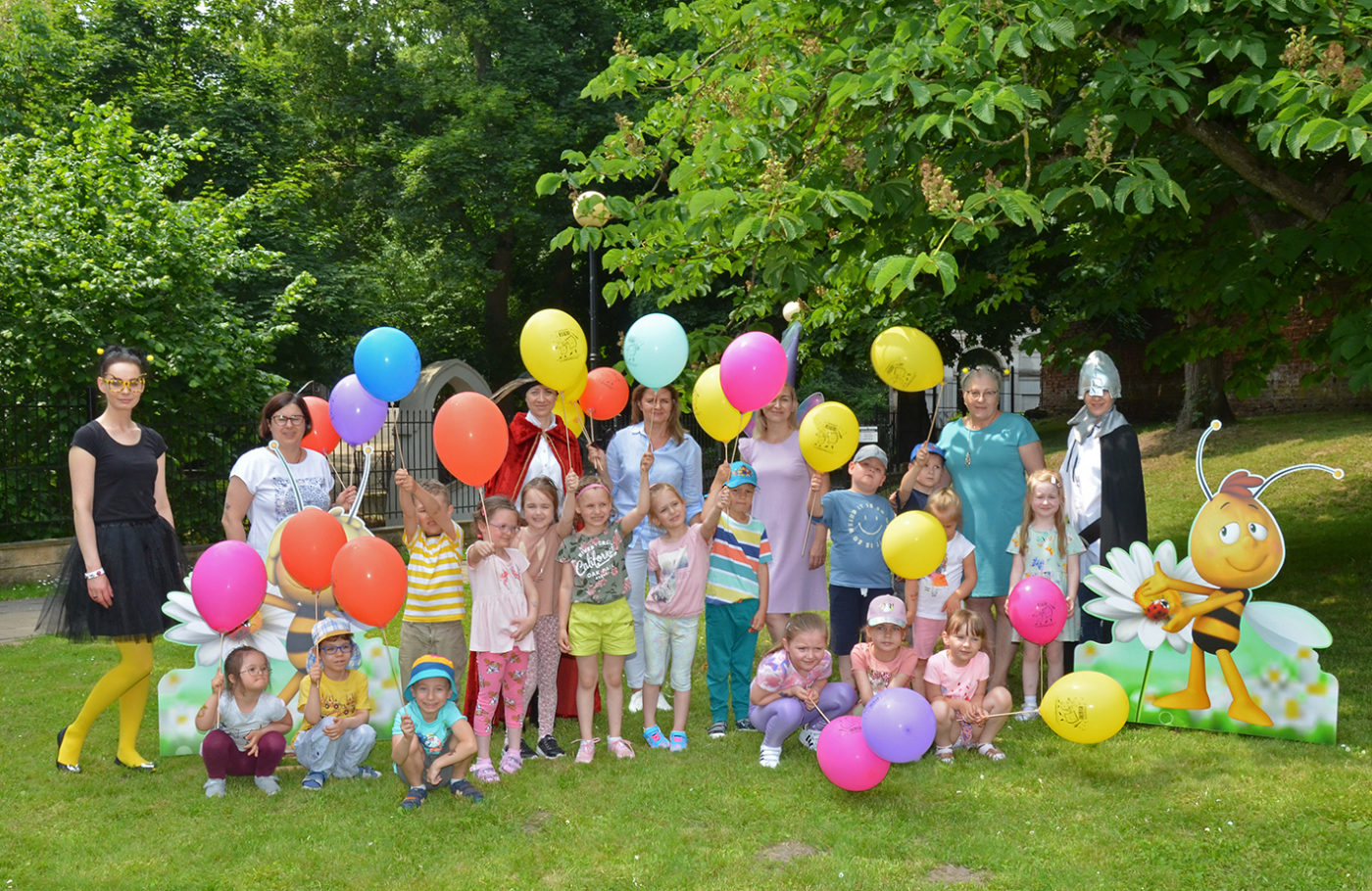 Grupa przedszkolaków pozuje do zdjęcia w otoczeniu osób przebranych za postaci z bajek. Unoszą do góry ręcę z kolorowymi balonami.